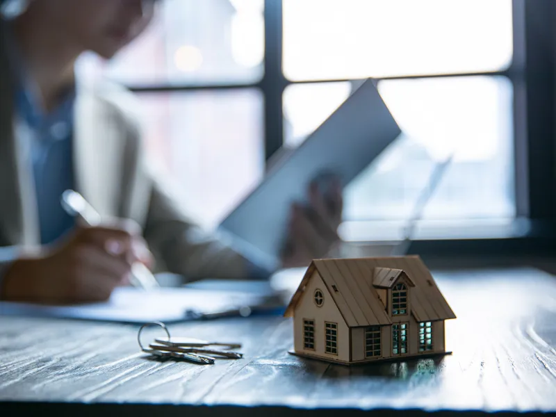 Model house and keys on a table with a person in the background reading a document and taking notes.